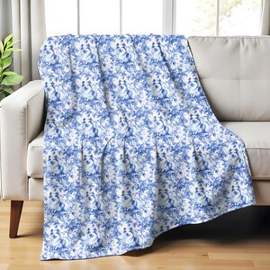 Puede incluir: Una manta estampada en azul y blanco sobre un sofá blanco. La manta presenta un diseño repetido de motivos florales y animales. El sofá tiene patas de madera y está colocado frente a una ventana.