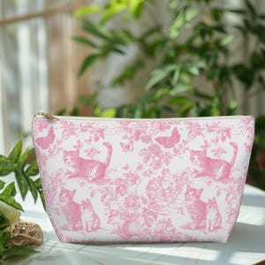 Op de afbeelding: Een witte make-uptas met een roze toile de Jouy-patroon met katten, vlinders en bloemmotieven. De tas heeft een gouden ritssluiting.