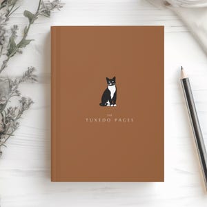 May include: A brown journal with the title "The Tuxedo Pages" and a black and white cat illustration. The journal is closed and has a pencil next to it. The journal is on a white surface.