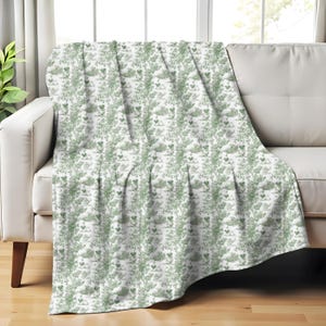 Op de afbeelding: Een witte sprei met een herhalend patroon van groene botanische illustraties, waaronder bladeren en kleine dieren, gedrapeerd over een witte bank. De sprei voegt een vleugje natuurlijke decoratie toe.