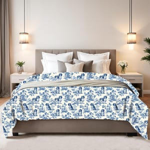 Puede incluir: Una manta estampada en blanco y azul con un diseño repetido de perros y elementos florales. La manta está colocada sobre una cama con un cabecero y almohadas de tonos neutros. La estética general es clásica y decorativa.