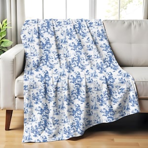 May include: A white throw blanket with a blue and white pattern of dogs and trees. The blanket is folded and draped over a white couch.