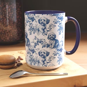 May include: A white ceramic mug with a blue floral and dog pattern. The mug has a blue interior and handle. The design features illustrations of dogs and floral elements in blue against a white background. A spoon and a pastry are in the foreground.