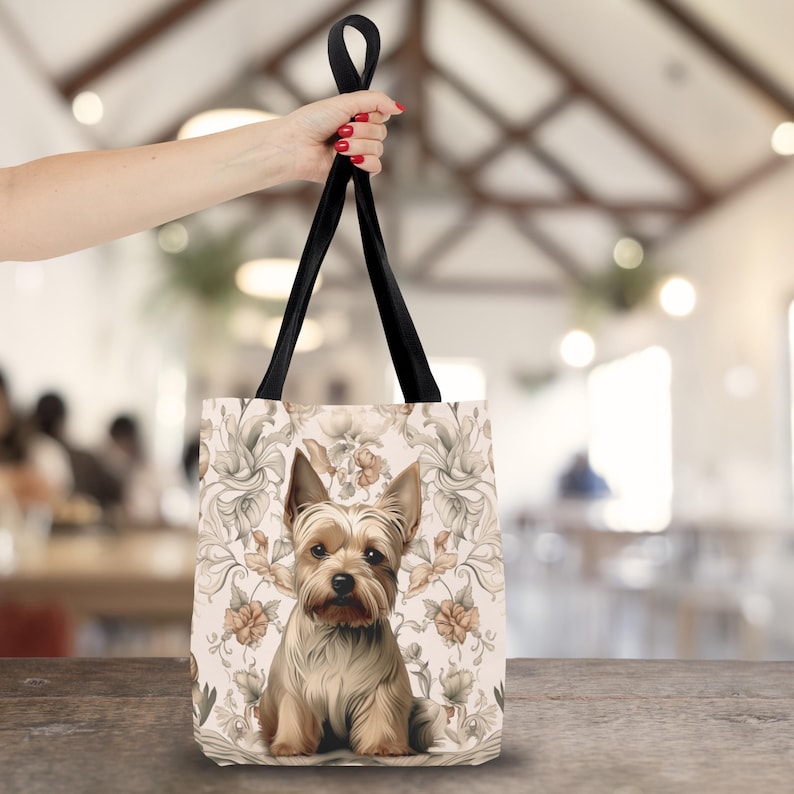 Pu&ograve; includere: Una borsa tote con un'illustrazione centrale di un cane Yorkshire Terrier marrone e beige. Lo sfondo della borsa presenta un motivo ripetuto di disegni floreali nei toni del beige e del marrone chiaro. La borsa ha due manici neri.