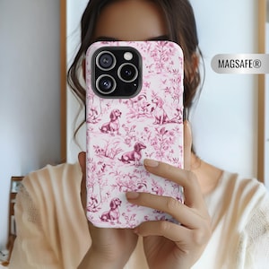 May include: A white phone case with a pink toile pattern featuring dachshunds and floral elements. The case has a MagSafe design and a triple-lens camera cutout.