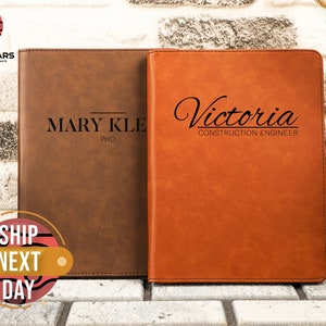May include: Two brown leather journals with personalized engravings. The left journal has the name "MARY KLE" and the title "PhD". The right journal has the name "Victoria" and the title "CONSTRUCTION ENGINEER".