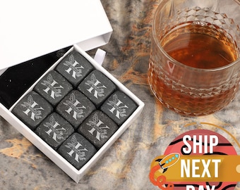 May include: A white box with a black velvet bag inside. The bag contains nine gray stone whiskey chilling cubes with a letter K engraved on each cube. The box is on a brown and gold textured surface.