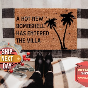 May include: A rectangular tan coir doormat featuring black text that reads A HOT NEW BOMBSHELL HAS ENTERED THE VILLA, accompanied by a black graphic illustration of two palm trees and several small birds flying in the background.
