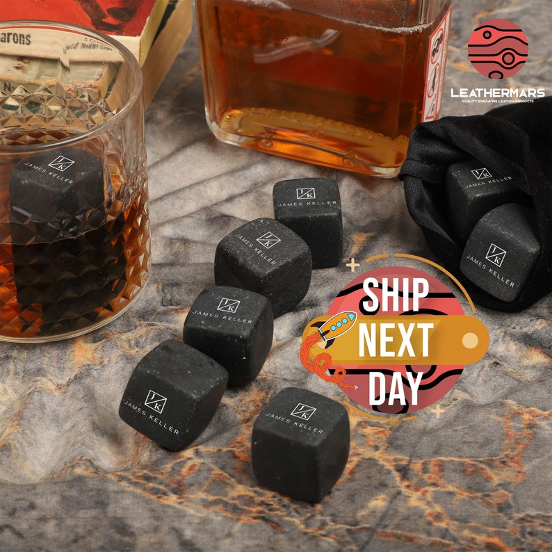 May include: Eight black square whiskey stones with the text "JAMES KELLER" on each stone. The stones are in a glass and on a table with a bottle of whiskey and a bag.