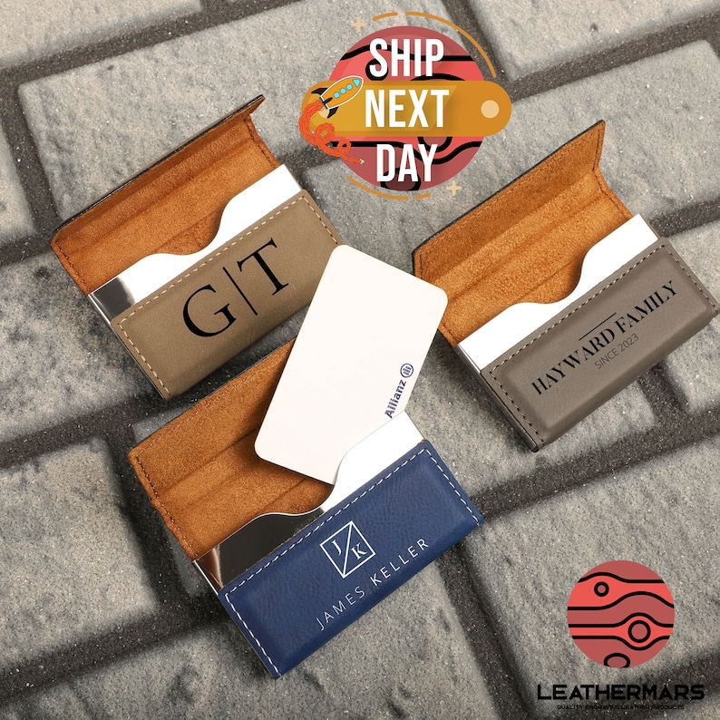 May include: Three leather business card holders with different personalized engravings. The holders are open to show the interior card slots and a business card inside each. The holders are brown, blue, and gray.