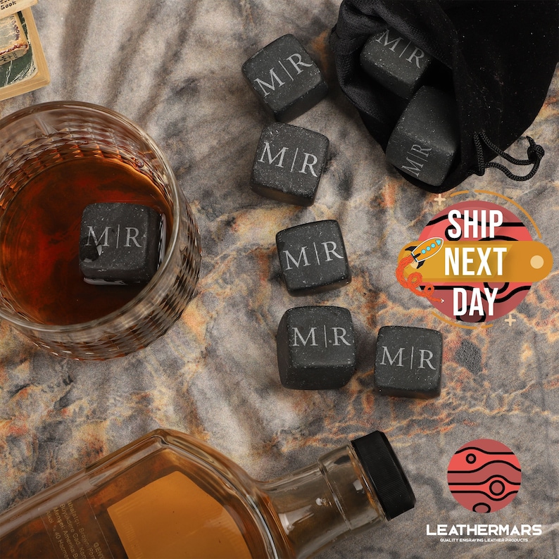 May include: Set of eight black granite whiskey stones with the initials "M/R" engraved on each stone. The stones are in a small black velvet bag and are next to a glass of whiskey and a bottle of whiskey.