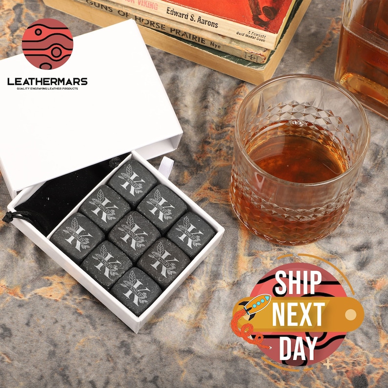 May include: A white box with a black velvet bag inside. The bag contains nine gray stone whiskey chilling cubes with a letter K engraved on each cube. The box is on a brown and gold textured surface.