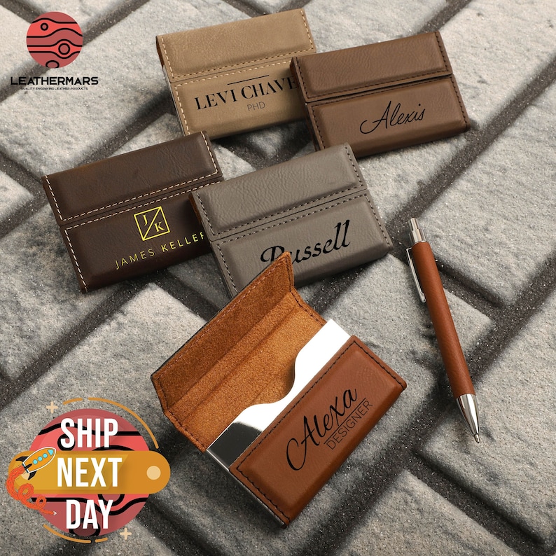 May include: Five different leather business card holders with engraved names. The holders are brown, gray, and tan. The holders are on a brick background. The text on the holders reads: "Levi Chave, PhD", "Alexis", "James Keller", "Russell", and "Alexa Designer".