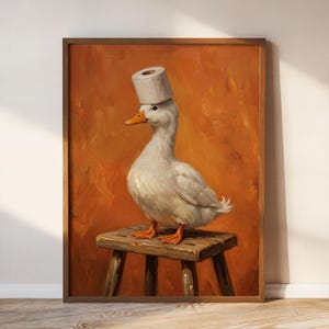 May include: A framed oil painting depicting a white duck with orange feet and a beak, wearing a roll of white toilet paper on its head like a hat. The duck is standing on a rustic wooden stool against a solid, textured, burnt orange background.