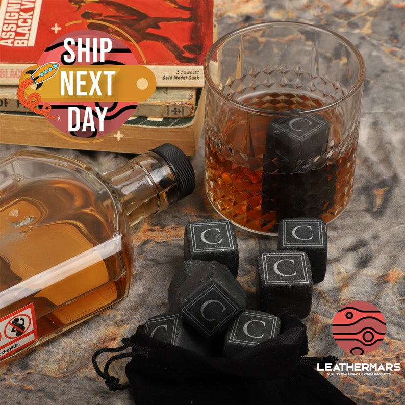 May include: Black whiskey stones with a letter C engraved on each side, in a black velvet bag. The stones are in a glass with whiskey and a bottle of whiskey is on the table.