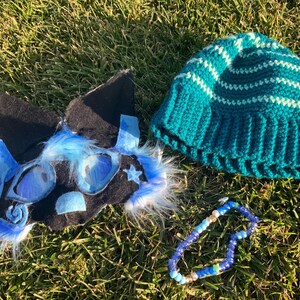 May include: A blue and black furry mask with a star and a swirl design, a teal and white striped crocheted beanie, and a blue beaded bracelet.