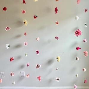 以下が含まれることがあります： ピンク、白、黄色の造花の花輪で飾られた壁。花は天井から吊り下げられており、ランダムなパターンで配置されています。