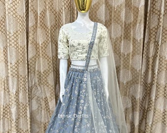 Op de afbeelding: Een lichtblauwe lehenga met ingewikkelde bloemenborduurwerk en een bijpassende dupatta. De outfit heeft een crèmekleurige top met korte mouwen en vergelijkbare versieringen. De tekst "Ethnic Outfits" is zichtbaar op de rok.