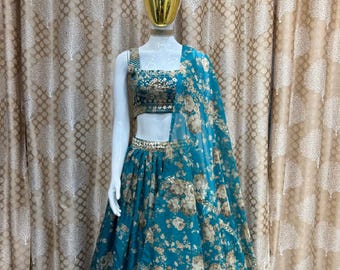 Op de afbeelding: Een tealblauwe en witte bloemenprint lehenga met een bijpassend mouwloos crop top. De top heeft een kraag met kralen en de rok heeft een gelaagd ontwerp. De outfit wordt getoond op een mannequin tegen een gepatroneerde achtergrond.
