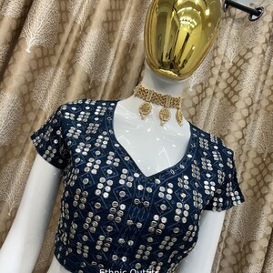 May include: A navy blue, short-sleeved top with a V-neck, heavily embellished with silver sequins in a geometric pattern. The text "Ethnic Outfits" is visible. A gold-colored necklace and earrings are also visible.