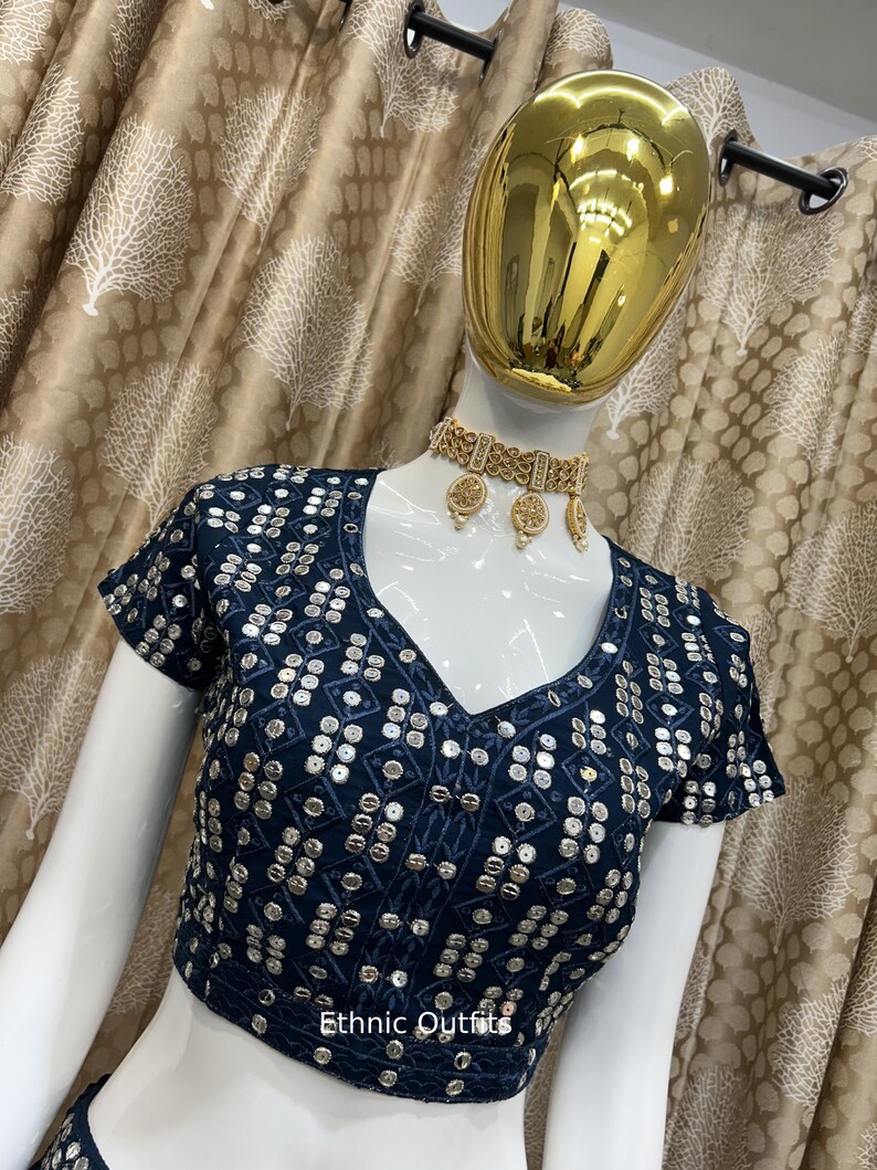 May include: A navy blue, short-sleeved top with a V-neck, heavily embellished with silver sequins in a geometric pattern. The text "Ethnic Outfits" is visible. A gold-colored necklace and earrings are also visible.