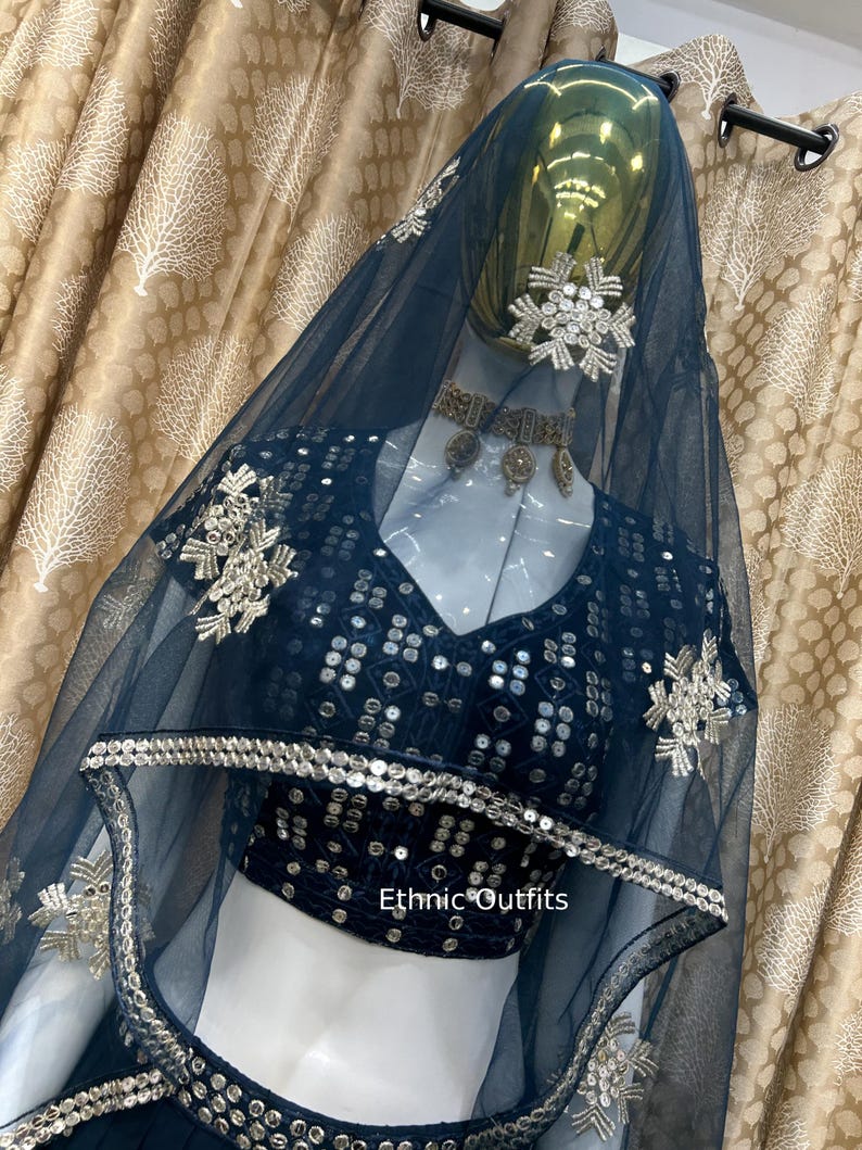 May include: A dark teal ethnic outfit, including a top and veil, displayed on a mannequin. The top is embellished with sequins and silver embroidery. The veil is sheer and adorned with decorative silver accents. The text "Ethnic Outfits" is visible.