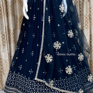 May include: A navy blue, embellished skirt and matching dupatta. The skirt features intricate silver embroidery along the hem and waist, with scattered decorative elements. The dupatta is sheer, with similar silver detailing and the text "Ethnic Outfits".