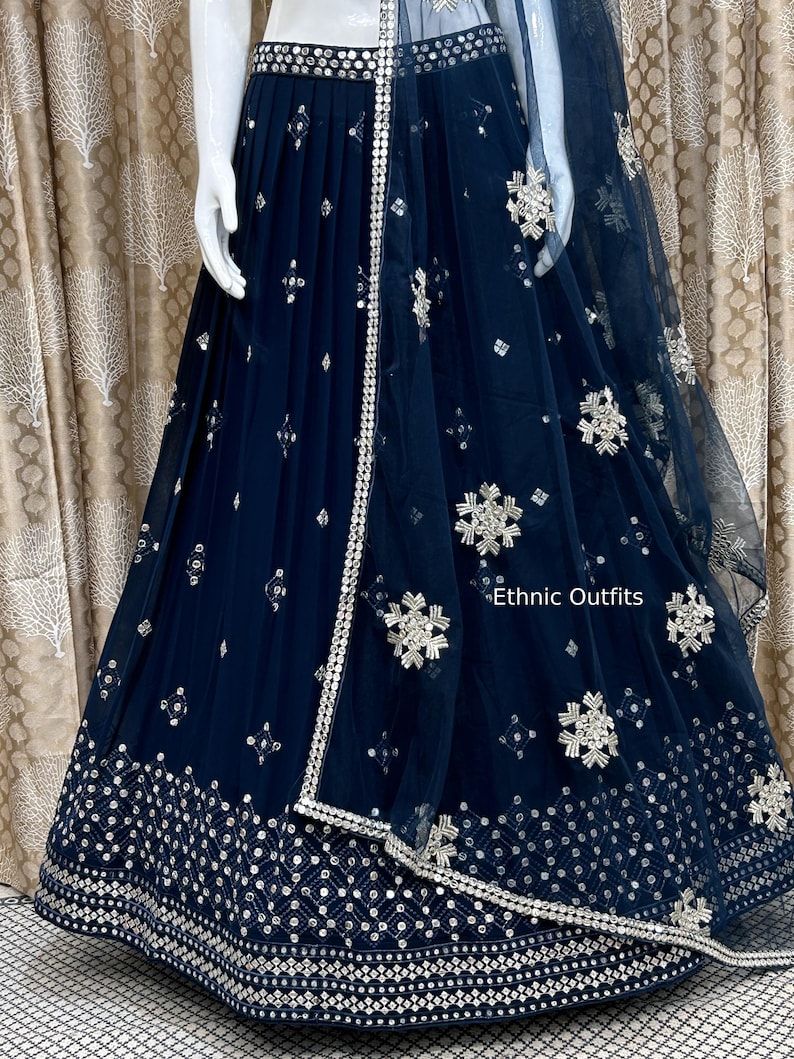 May include: A navy blue, embellished skirt and matching dupatta. The skirt features intricate silver embroidery along the hem and waist, with scattered decorative elements. The dupatta is sheer, with similar silver detailing and the text "Ethnic Outfits".