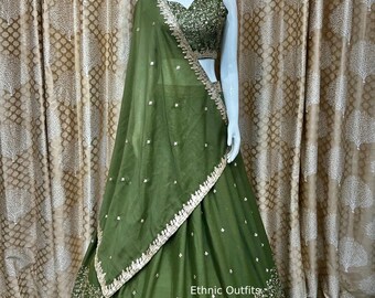 Op de afbeelding: Een groene en gouden geborduurde lehenga met een bijpassend mouwloos blouse en een transparante dupatta. De lehenga heeft een gouden rand en ingewikkelde bloemmotieven. De outfit wordt getoond op een mannequin tegen een bruine, gepatroneerde achtergrond.