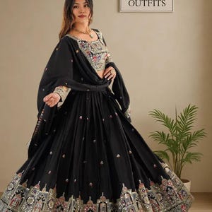 May include: Black ethnic outfit with intricate embroidery and embellishments. The outfit includes a top, a flowing skirt, and a matching scarf. The top features detailed embroidery around the neckline and sleeves. The skirt has a decorative border. The sign in the background says "ETHNIC OUTFITS".