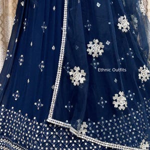 May include: Navy blue formal dress with intricate silver embroidery and embellishments. The dress features a detailed border and delicate floral patterns. The text "Ethnic Outfits" is visible on the dress.