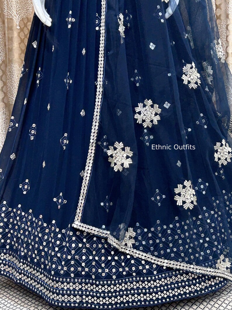 May include: Navy blue formal dress with intricate silver embroidery and embellishments. The dress features a detailed border and delicate floral patterns. The text "Ethnic Outfits" is visible on the dress.