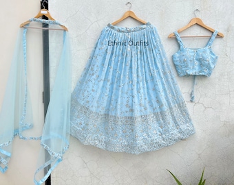 Puede incluir: Un conjunto de tres piezas azul claro, que incluye falda, top y chal, todos adornados con detalles plateados. La falda y el top están adornados con pequeños detalles reflectantes. El conjunto se muestra en perchas de madera sobre un fondo neutro. El texto "Ethnic Outfits" es visible.