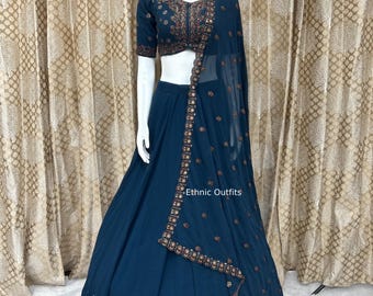 May include: A teal-colored ethnic outfit displayed on a mannequin. The outfit includes a short-sleeved top with intricate embroidery, a flowing skirt, and a matching sheer scarf. The scarf and top are adorned with detailed embellishments. The text "Ethnic Outfits" is visible.