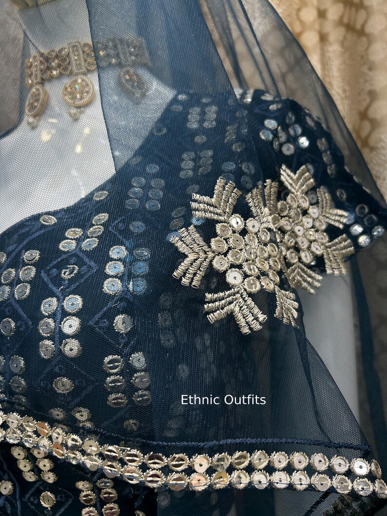 May include: Close-up of a navy blue ethnic outfit with intricate silver embroidery and sequin detailing. The outfit features a sheer overlay and the text "Ethnic Outfits". The design includes floral and geometric patterns.