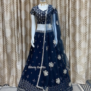 May include: A navy blue lehenga choli set on a mannequin. The set includes a short-sleeved top and a long skirt, both embellished with silver sequins and floral patterns. A sheer dupatta with matching embellishments completes the ensemble. The text "Ethnic Outfits" is visible.
