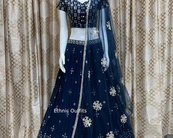 May include: A navy blue lehenga choli set displayed on a mannequin. The outfit features a short-sleeved top and a long skirt, both adorned with silver sequins and floral designs. A sheer dupatta with similar embellishments complements the set. The text "Ethnic Outfits" is visible.