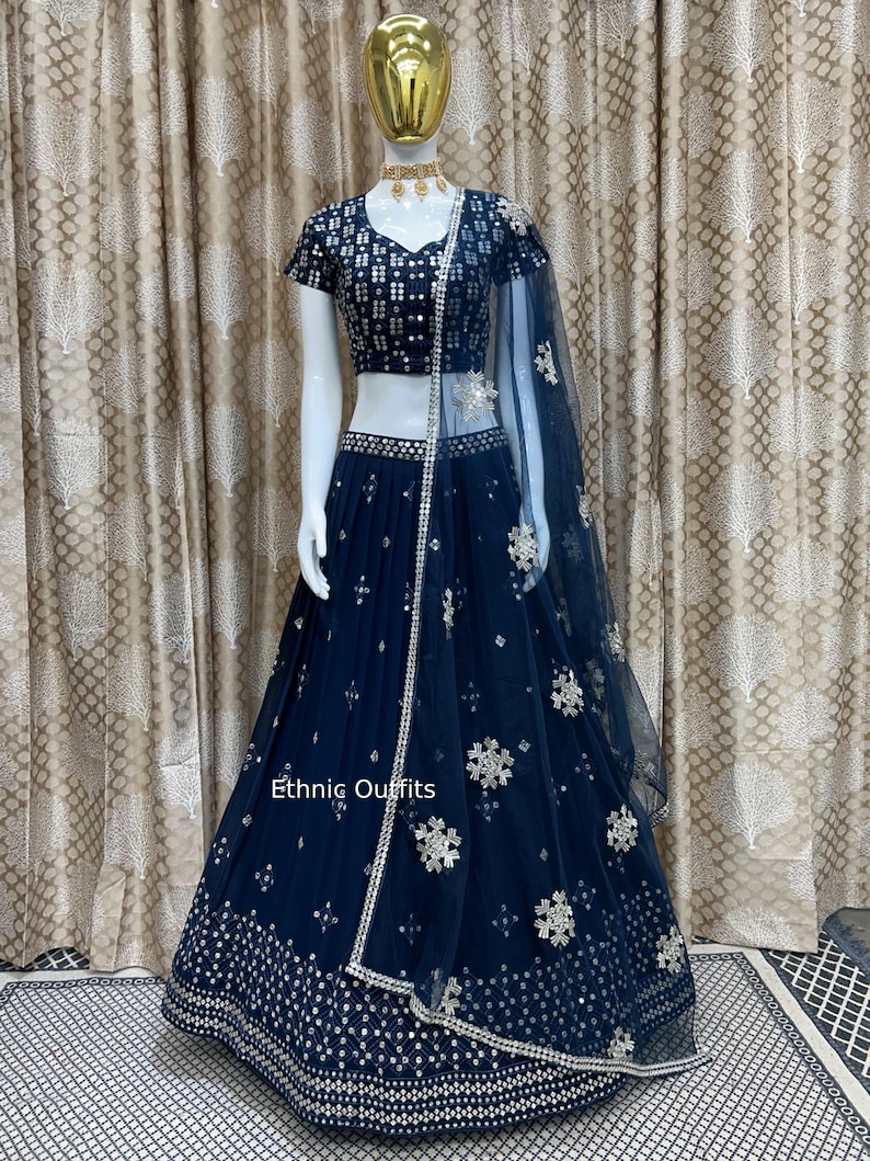 May include: A navy blue lehenga choli set on a mannequin. The set includes a short-sleeved top and a long skirt, both embellished with silver sequins and floral patterns. A sheer dupatta with matching embellishments completes the ensemble. The text "Ethnic Outfits" is visible.