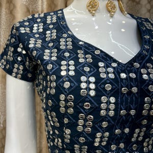 May include: A navy blue, short-sleeved top with a V-neck, adorned with silver sequins in a geometric pattern. The top is part of a set, paired with a gold-toned choker necklace and matching earrings. The text "Ethnic Outfits" is visible.