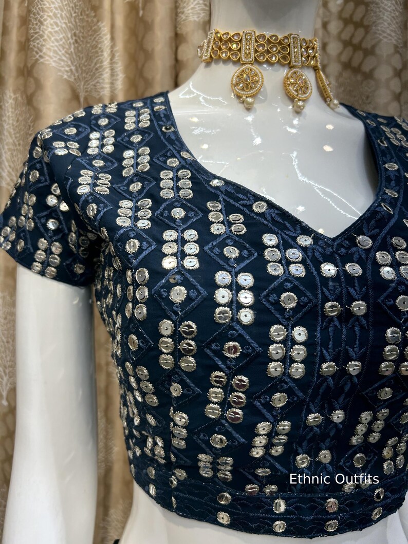 May include: A navy blue, short-sleeved top with a V-neck, adorned with silver sequins in a geometric pattern. The top is part of a set, paired with a gold-toned choker necklace and matching earrings. The text "Ethnic Outfits" is visible.