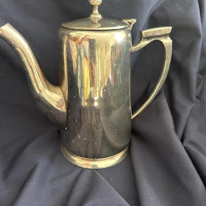 Puede incluir: Una cafetera plateada con un pico curvo y un asa. La cafetera tiene una tapa con un pequeño pomo decorativo en la parte superior. La cafetera está sobre una superficie azul oscuro.