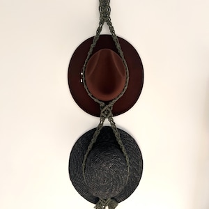 May include: A macramé hat holder with a wooden ring at the top, displaying a brown fedora and a black sun hat. The dark green macramé suspends the hats against a white wall.
