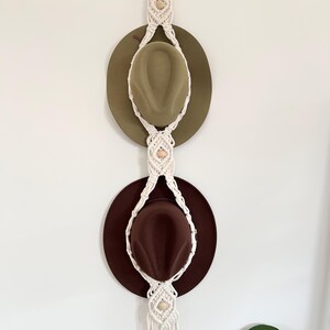May include: A macramé hat rack with two hats. The rack is made of cream-coloured woven cotton and features wooden beads. The hats are olive green and brown. The rack is hanging on a white wall.