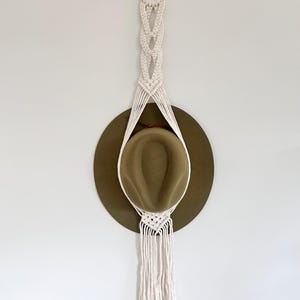 May include: A cream-coloured macrame hat holder with a wooden ring and woven pattern. An olive green hat is suspended within the holder, showcasing a bohemian style. The hat holder is mounted on a white wall.