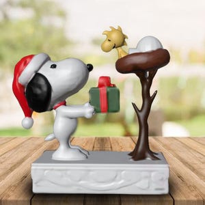 May include: A white ceramic figurine of Snoopy wearing a red Santa hat, holding a green and red gift box.  Snoopy is standing on a white base with a brown tree and a yellow bird figurine on top.