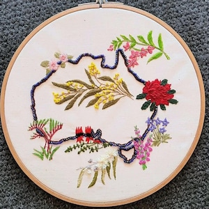 May include: Embroidered artwork in a wooden hoop, depicting the Australian continent. The outline is formed with dark blue beads, and the interior is filled with colorful floral embroidery. The flowers are in shades of red, pink, yellow, and purple, on a cream background.