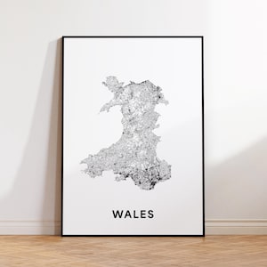 May include: A framed print displaying a detailed black and white map of Wales. The map shows the country's shape and internal features. The word "WALES" is printed in bold, black letters at the bottom of the print, below the map.