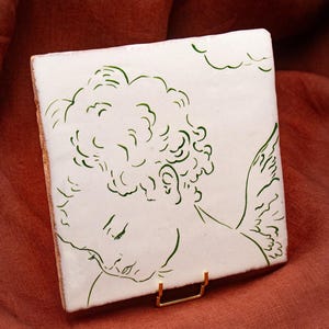 Peut inclure: Un carreau de céramique blanc carré orné d'un dessin au trait vert représentant un chérubin aux cheveux bouclés et aux ailes. Le carreau est soutenu par un petit support en métal doré, mettant en valeur un design artistique épuré.