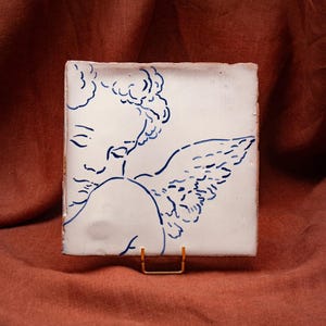 Peut inclure: Un carreau en céramique blanc de forme carrée orné d'une illustration au trait bleu représentant un chérubin aux cheveux bouclés avec une aile. Le carreau est présenté sur un petit support en métal doré, révélant une surface lisse.
