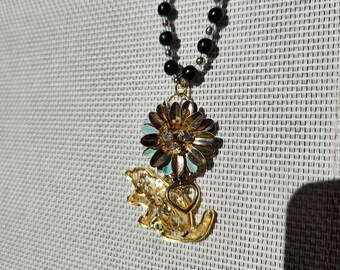 May include: A gold necklace with a black bead chain and a pendant featuring a gold flower with blue and white accents, a gold angel with a heart charm, and a gold key.